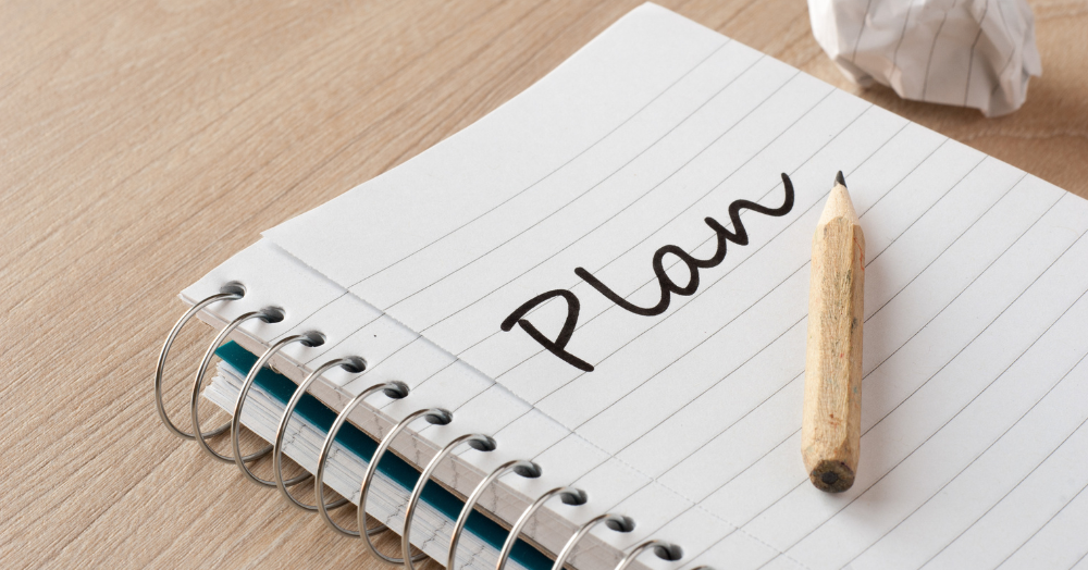 Slika prikazuje beležko z napisano besedo “Plan” ter svinčnik in zmečkan list papirja v ozadju