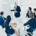 Skupina poslovnih ljudi na sestanku, kjer vodja predstavitve kaže graf na beli tabli. Sestanek poteka v moderni pisarni s steklenimi okni v ozadju.