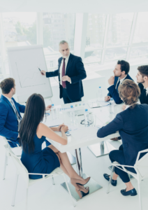 Skupina poslovnih ljudi na sestanku, kjer vodja predstavitve kaže graf na beli tabli. Sestanek poteka v moderni pisarni s steklenimi okni v ozadju.